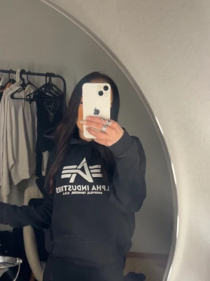 Svart hoodie från Alpha Industries - Svart hoodie från Alpha Industries med stor vit logga och text på bröstet. Klassisk modell med huva och känguruficka framtill. Perfekt för en avslappnad och cool streetwear-look.