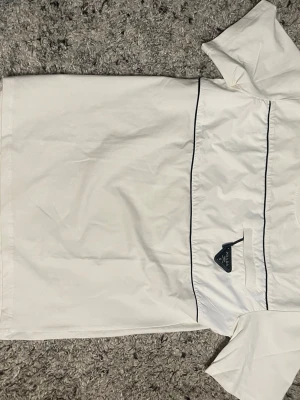 Vit t-shirt från Prada med ficka - Snygg vit t-shirt från Prada med svart kantband och en liten bröstficka. På fickan sitter den ikoniska svarta Prada-loggan i metall. T-shirten har en clean och modern look, perfekt för dig som gillar exklusiva detaljer och stilren design.