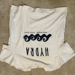 Vit HYDRA t-shirt med blått tryck - Vit t-shirt från HYDRA med blått vågmönster och texten 'Hydra' på bröstet. Klassisk passform med korta ärmar och rund halsringning. Tillverkad i mjuk bomull, perfekt för en chill och stilren look.