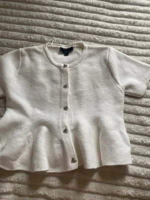 Vit kortärmad kofta från Zara - Supersöt vit kofta från Zara med korta ärmar och hjärtformade knappar framtill. Koftan har en lätt utsvängd passform och är stickad i ett mjukt material. Perfekt att slänga över en topp för en söt look.