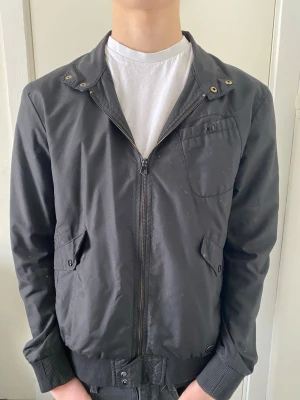 Svart bomberjacka med dragkedja - Stilren svart bomberjacka med dragkedja framtill, ribbade muddar och krage med tryckknappar. Jackan har två snedställda fickor med lock och en bröstficka. Perfekt för dig som gillar en clean och enkel look. Den har dessutom ett hål i armhålan inuti jackan. Det syns inte alls när man har på sig den. Annars är det inget fel på jackan alls.