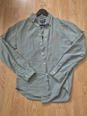 Rutig skjorta från Polo Ralph Lauren - Klassisk rutig skjorta från Polo Ralph Lauren i grönt och vitt med lång ärm och knappar framtill. Skjortan har button-down krage och den ikoniska röda logotypen broderad på bröstet. Perfekt för en avslappnad men stilsäker look.