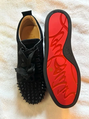 Svarta spikes-sneakers Christian Louboutin - Unika svarta sneakers från Christian Louboutin med mattsvarta spikes på tån och klassisk röd sula. Skorna har svarta skosnören och är tillverkade i mocka. 