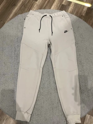 Beige mjukisbyxor från Nike - Snygga beige mjukisbyxor från Nike med svarta dragsnören och logga på vänster sida. Byxorna har en slim fit-modell, muddar vid bensluten och praktiska fickor. Perfekta för en avslappnad och sportig stil.