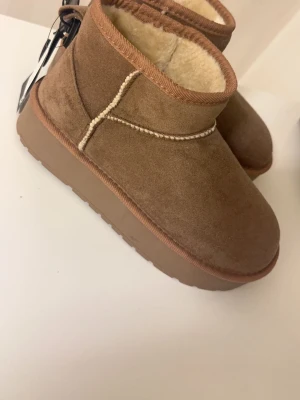 Hel nya Bruna boots med foder och platåsula - Mysiga bruna boots med mjukt foder och kraftig platåsula. Utsidan är i mockaliknande material och insidan är fodrad med beige, fluffigt tyg. Snygga detaljer med synliga sömmar och låg skaftmodell. Perfekta för kalla dagar.bootsen är oanvända helt nya