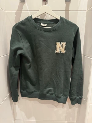 Mörkgrön sweatshirt med N-patch från Kappahl - Mörkgrön sweatshirt från Kappahl i mjukt material med rund halsringning och ribbade muddar. På bröstet sitter en beige patch med bokstaven N i college-stil. Perfekt för en avslappnad och trendig look.