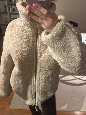 Beige teddyjacka med dragkedja - Mysig beige teddyjacka med hög krage och dragkedja framtill. Jackan har en fluffig, fårullsliknande yta och avslappnad passform. Perfekt för kalla dagar när du vill vara både varm och snygg. Den funkar även bra nu till våren!!