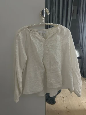 Vit blus från Zara - Säljer en vit blus från Zara med långärmad design och rundad halsringning. Blusen har ett strukturerat tyg med diskreta detaljer framtill och lätt puffade ärmar. Perfekt för dig som gillar en enkel men ändå trendig look.