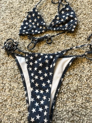 Svart bikini med stjärnor från MNG - Snygg svart bikini från MNG med vita stjärnor över hela tyget. Klassisk trekantsmodell på toppen och knyt i sidorna på underdelen. Perfekt för stranden eller poolen. Materialet är mjukt och stretchigt för skön passform.