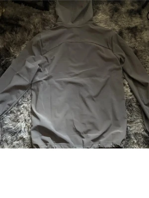 Grå vindjacka från Under Armour - Snygg grå vindjacka från Under Armour med huva och dragkedja framtill. Jackan har två sidofickor och en diskret logga på bröstet. Tillverkad i ett lätt och vindtätt material som passar perfekt för blåsiga dagar.