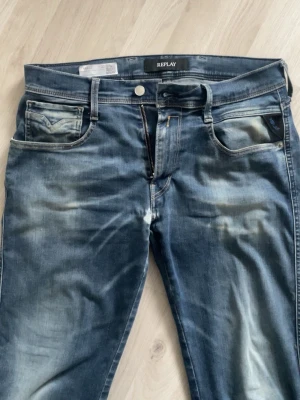 Blå jeans från Replay - Säljer ett par blå jeans från Replay med tvättade detaljer. Klassisk femficksmodell med normal midja och raka ben. Jeansen har en cool, använd look och är perfekta för en avslappnad stil. Jeansen har även ett par slitningar men bortsett från det är skicket felfritt!