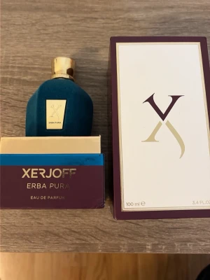 Xerjoff Erba Pura Eau de Parfum - Lyxig parfym från Xerjoff, Erba Pura, i en elegant blå flaska med gulddetaljer och tillhörande ask. Flaskan har en mjuk, sammetsliknande yta och guldfärgat lock. Förpackningen är stilren med guld- och lila detaljer. Volym: 70ml.