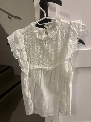 Vit blus med spetsdetaljer - Söt vit blus med broderade spetsdetaljer framtill och volangprydda ärmar. Blusen har en rund halsring sopps med knapp i nacken och romantiska detaljer läng following axlarna. 