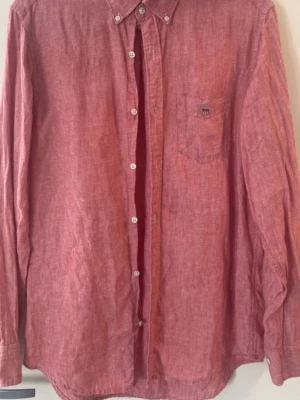 Röd linneskjorta från GANT - Säljer en snygg röd skjorta från GANT i linne med klassisk krage och knappar hela vägen. Skjortan har en bröstficka med broderad GANT-logga och emblem. Perfekt för dig som gillar stilrena och bekväma plagg.