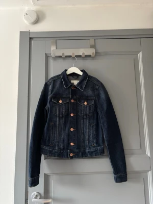 Jeansjacka - Snygg mörkblå jeansjacka med kopparfärgade knappar och två bröstfickor med lock. Jackan har en tidlös design med raka ärmar och justerbar midja baktill. Perfekt att slänga över en hoodie eller t-shirt för en chill look.