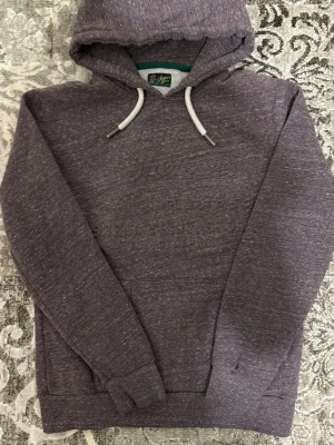 Superdry Hoodie - Mysig hoodie i melerad grå färg från Superdry med broderad logga på bröstet. Tröjan har huva med vita dragsnören och ribbade muddar vid ärmslut och nederkant. Perfekt för en avslappnad och cool stil.