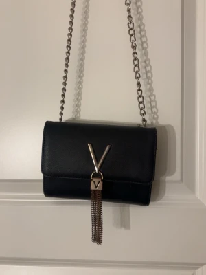 Svart axelväska från Valentino - Stilren svart axelväska från Valentino med silverfärgad kedja och metall-detalj i form av ett V på framsidan. Väskan har en elegant tassel av kedjor och är tillverkad i ett strukturerat syntetmaterial. Perfekt för dig som gillar trendiga accessoarer.