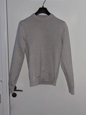 Beige stickad tröja från Dobber - En stilren beige stickad tröja från Dobber i storlek S. Tröjan har rund halsringning, ribbade muddar vid ärmslut och nederkant samt en klassisk rak passform. Perfekt att matcha med jeans eller kjol för en enkel och snygg look.
