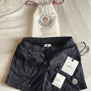 Moncler shorts - Snygga svarta shorts från Moncler med broderad logotyp. De har en praktisk dragkedja på baksidan och snörning i midjan för justerbar passform. Perfekta för sommaren.