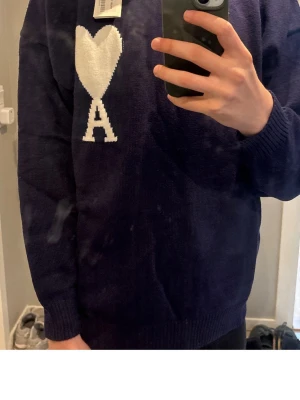 Mörkblå stickad tröja Ami Paris - Säljer en mörkblå stickad tröja från Ami Paris med deras ikoniska vita hjärta och bokstaven A på bröstet. Tröjan har rund halsringning och ribbade muddar vid ärmslut och nederkant. Perfekt för dig som gillar stilrena detaljer.