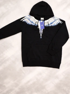Svart hoodie Marcelo Burlon med vingar - Svart hoodie från Marcelo Burlon County of Milan med ikoniskt vingtryck i vitt och blått över axlar och bröst. Klassisk känguruficka framtill och justerbar huva. Tillverkad i mjuk bomull för skön känsla och stilren look.
