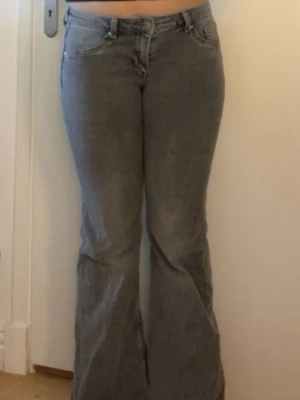 Grå bootcut jeans med låg midja - Säljer ett par gråa bootcut jeans med låg midja och klassiska fem fickor. Jeansen har en lätt tvättad look och stängs med dragkedja och knapp. Perfekta för en avslappnad och trendig stil.