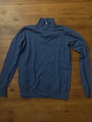 Blå stickad tröja med halv dragkedja - Mörkblå stickad tröja med hög krage och halv dragkedja framtill. Tröjan har långa ärmar och ribbstickade muddar vid ärmslut och nederkant. Perfekt för lager-på-lager och en stilren look.