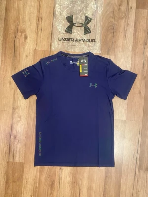 Blå Under Armour t-shirt - Snygg blå t-shirt från Under Armour med logga på bröstet och tryck på ärmen. Perfekt för träning med sin sportiga design och bekväma passform. Gjord i lätt och funktionellt material som andas. Klassisk rund hals och korta ärmar.