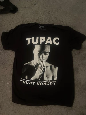 Svart 2PAC T-shirt med tryck - Säljer en svart T-shirt från 2PAC med stort tryck av Tupac och texten 'Trust Nobody' på framsidan. Klassisk passform och rund hals. Perfekt för dig som gillar streetwear och ikoniska prints.