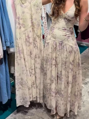 Adoore klänning - Adoore Toulouse klänning. långklänning i krämfärg/gul med lila och rosa blommigt mönster. Klänningen har v-ringning, markerad midja och är ärmlös. Perfekt för sommarens alla tillfällen och ger en bohemisk känsla.