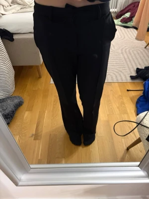 Svarta kostymbyxor med bootcut - Säljer ett par svarta kostymbyxor med bootcut och pressveck. Byxorna har fickor fram och hällor för bälte. Snygg passform som sitter bra över höften och släpper vid benen. Perfekta för dig som vill ha en klassisk och stilren look.