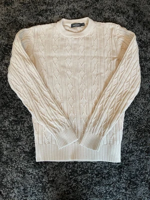 Kabelstickad offwhite tröja Dressmann - Kabelstickad tröja från Dressmann i en snygg offwhite färg. Tröjan har rund halsringning, ribbade muddar och ett klassiskt stickat mönster. Perfekt för lager-på-lager och passar till många olika stilar.