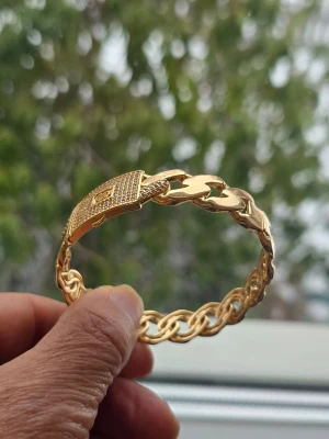 Guldigt kedjearmband med låsdetalj - Snyggt armband i guldfärgad metall med en chunky kedjedesign och en unik låsdetalj framtill. Armbandet har en flätad struktur på ena sidan och bredare länkar på andra, vilket ger en cool och modern look. Perfekt accessoar för att lyfta din outfit. Kopar 