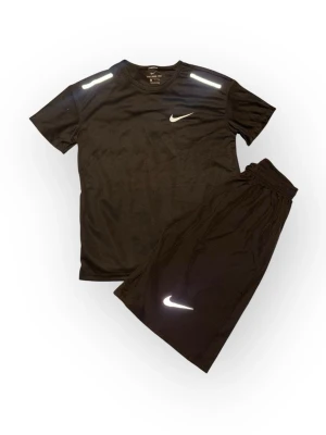 Svart träningsset från Nike - Snyggt svart träningsset från Nike med t-shirt och shorts. Båda plaggen har vita Nike-loggor och reflekterande detaljer på axlar och ben. Tillverkade i lätt och snabbtorkande funktionsmaterial som passar perfekt till träning och sport.