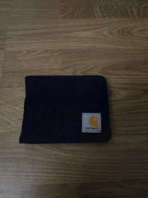 Svart manchesterplånbok Carhartt - Snygg svart plånbok i manchestertyg från Carhartt. Plånboken har flera kortfack, myntfack med knapp och klassisk Carhartt-logga i gult och vitt på framsidan. Perfekt för dig som gillar streetwear och vill ha något stilrent och praktiskt.