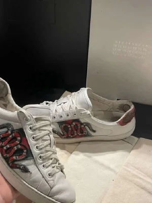 Gucci Ace sneakers med orm - Snygga vita Gucci Ace sneakers i läder med ikonisk broderad röd och svart orm på sidan. Skorna har gröna och röda detaljer på hälen och klassisk låg siluett. Perfekta för dig som vill sticka ut med en lyxig och trendig look. Enda stora defekt är sulan under skon som är sliten, skon är även overall ganska använd 