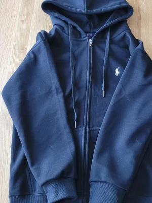 raulph lauren tröja - Klassisk svart hoodie från Polo Ralph Lauren med dragkedja framtill och vit broderad logga på bröstet. Hoodien har huva med dragsko, ribbade muddar och fickor på sidorna. Perfekt för en chill och stilren look.