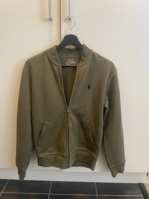 Olivgrön zip hoodie från Polo Ralph Lauren - Snygg olivgrön zip från Polo Ralph Lauren med dragkedja framtill och två fickor. Klassisk logga broderad på bröstet. Tillverkad i mjukt bomullsmaterial. Perfekt för en avslappnad och stilren look.