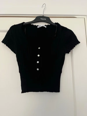 Svart croppad topp  - Snygg svart croppad topp från Zara med korta ärmar, knappar framtill och söt knytning i midjan. Toppen har en v-ringad, vågig kant och diskreta håldetaljer upptill. Perfekt till jeans eller kjol för en trendig look.