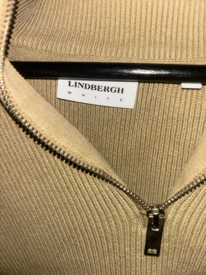 Beige stickad halvzip från Lindbergh - Stickad tröja i beige från Lindbergh med halvzip och hög krage. Tröjan har ribbad struktur och långa ärmar, perfekt för lager på lager. Enkel och stilren design som funkar till det mesta. Använd lite nytt skick storlek M
