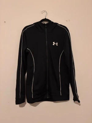 Tracksuit Under Armour - Svart hoodie med dragkedja från Under Armour. Hoodien har vita detaljer längs ärmar och sidor samt en vit logga på bröstet. Tillverkad i ett mjukt och stretchigt material, perfekt för träning eller chill. Klassisk passform med huva och långa ärmar.