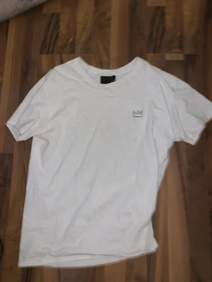 Vit t-shirt Hov1 Vi Dansar Vidare - Vit t-shirt från Hov1 med liten krona tryckt på bröstet och färgglatt turnétryck på ryggen med texten 'Vi Dansar Vidare I Livet' och turnédatum. Klassisk passform och rund hals. Perfekt för dig som gillar svensk musik och streetstyle.