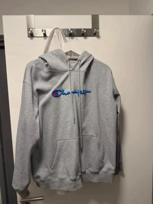 Grå hoodie från Champion M/XL - VERKLIGEN INTE XL. Mer M/S. Snygg grå hoodie från Champion med blå och röd broderad logga på bröstet. Klassisk känguruficka framtill, justerbar huva med dragsko och mjukt material på insidan. Perfekt för en chill och sportig stil.