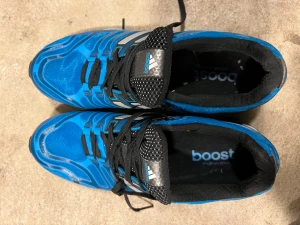 Blå Adidas Boost sneakers - Snygga blå sneakers från Adidas med Boost-sula och svarta detaljer. Skorna har silvriga ränder på sidorna, svart mesh vid tungan och svarta skosnören. Ovandelen är i syntetmaterial och mesh för bra andning. Perfekta för dig som gillar sportig stil.