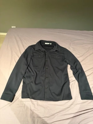 Svart overshirt med fickor - Stilren marinblå overshirt med knäppning framtill och två bröstfickor med lock. En perfekt overshirt för lager på lager. Knappt använd, skulle gissa på runt 3-5 gånger. Storleken är M. Säljs för 200kr kan gå ner i pris vid snabb och smidig affär. Säljer bara genom PostNord. 