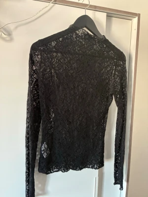 Svart spetsblus med lång ärm - Säljer en svart blus i spets med långärmad design. Toppen är genombruten och har en elegant, slim passform som framhäver siluetten. Perfekt för dig som gillar en stilren och trendig look med lite edge.