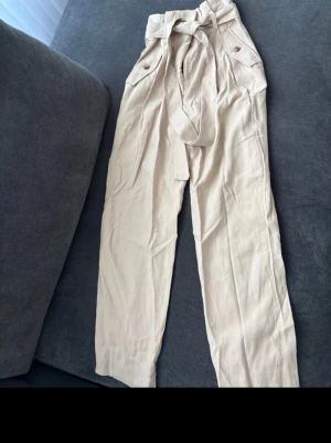 Beiga linnebyxor med knytskärp H&M - Snygga beiga linnebyxor från H&M med hög midja, knytskärp och fickor med knappdetaljer. Byxorna har raka ben och är tillverkade i ett luftigt linnematerial som passar perfekt för en avslappnad stil.