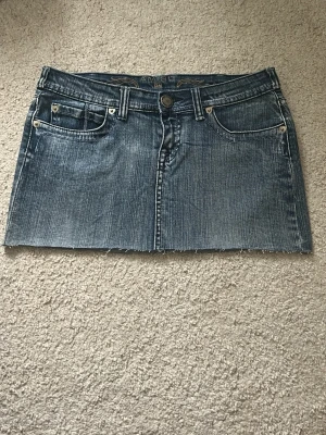 Blå jeanskjol med nitar från Amisu - Snygg blå jeanskjol från Amisu med rå kant nertill och dekorativa nitar på bakfickorna. Klassisk femficksmodell med bälteshällor och knappstängning. Perfekt för dig som gillar en trendig och avslappnad stil.