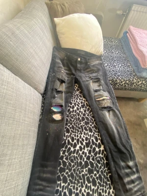 Svarta slitna jeans med hål - Coola svarta jeans med flera slitningar och hål på framsidan, samt färgstänk och regnbågsdetaljer under vissa hål. Smal passform och klassisk femficksdesign. Perfekta för en edgy streetstyle-look.