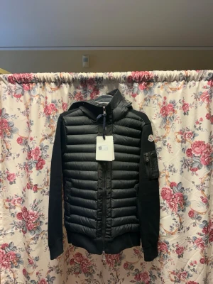 Padded zip up från Moncler - Svart pufferjacka från Moncler med quiltad front, stickade ärmar och huva. Jackan har dragkedja, två fickor fram och en ärmficka med Moncler-logga. Perfekt för kalla dagar och snygg streetstil.